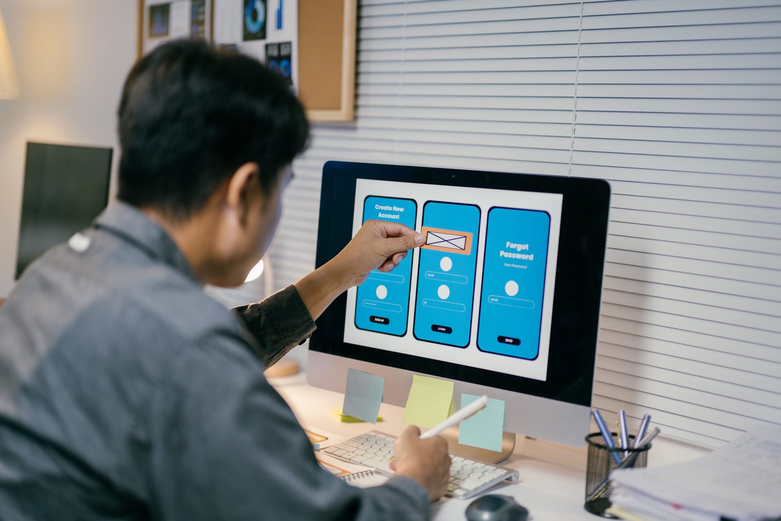 Ui developer is working on a user interface for a mobile application, using a wireframe kit displayed on his computer screen. He is holding a pen in his other hand and seems to be taking notes