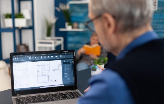 Overshoulder shot of senior architect working on laptop Overshoulder shot of senior architect working on laptop to finish a project. Experienced construction engineer designer looking at house blueprints on laptop computer from home office.