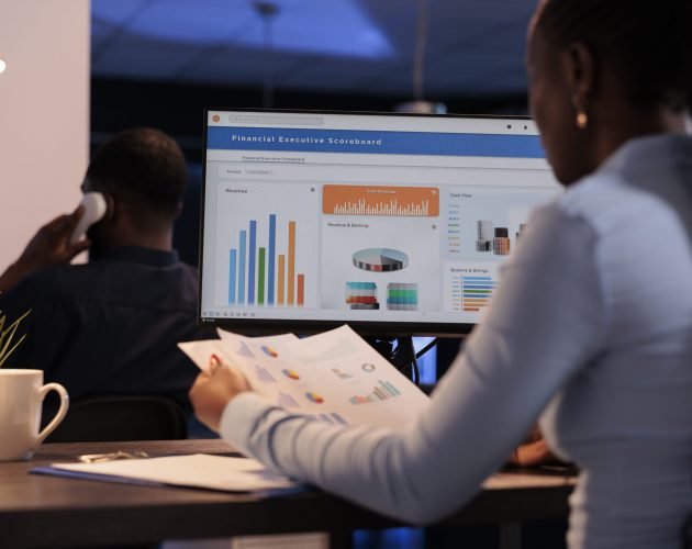 Young businesswoman working with statistics charts on computer monitor, analyzing company data on paperwork. Using online information to create research report before deadline, at night after hours.